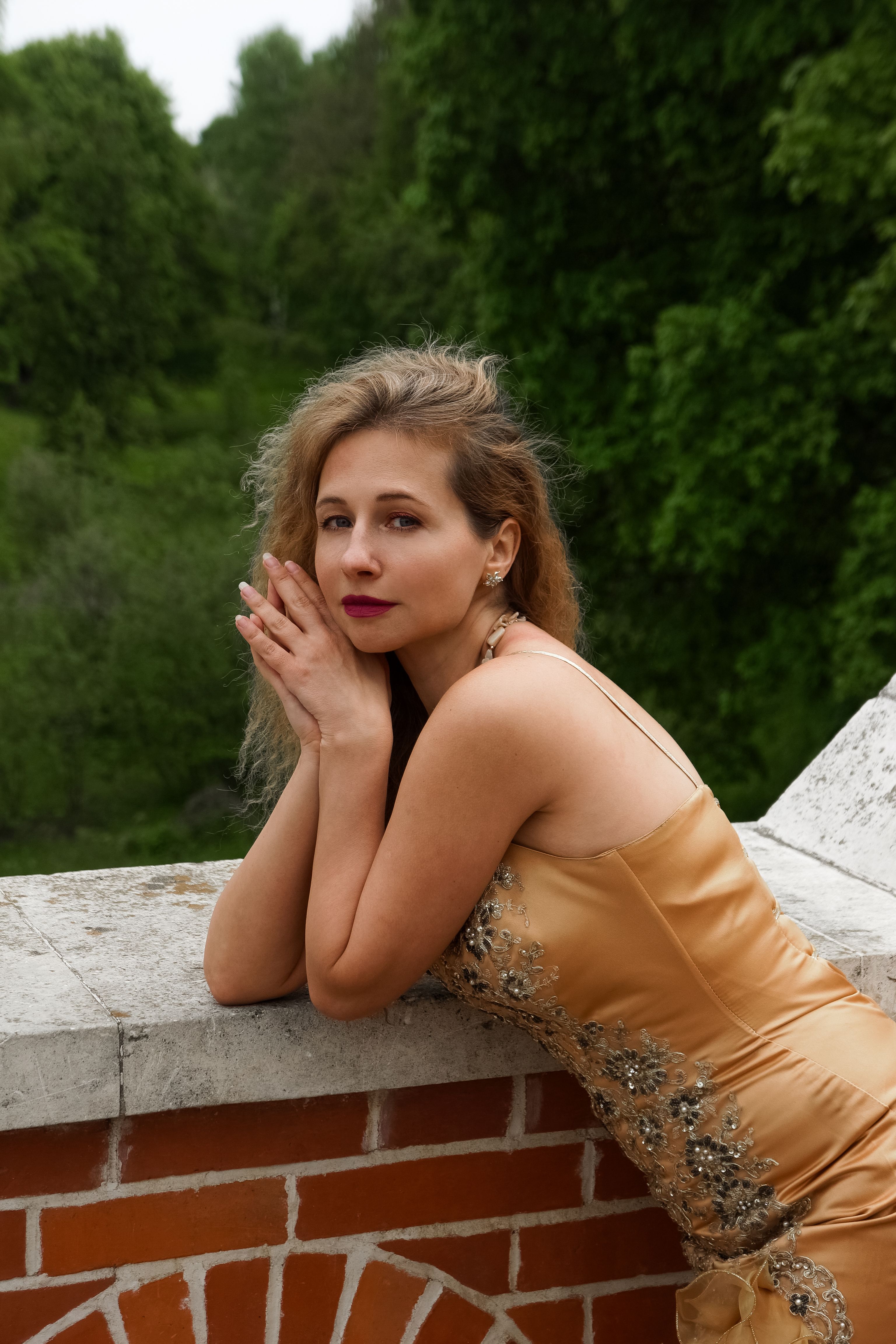 A woman in a gold dress poses gracefully against a brick wall in a lush outdoor setting.