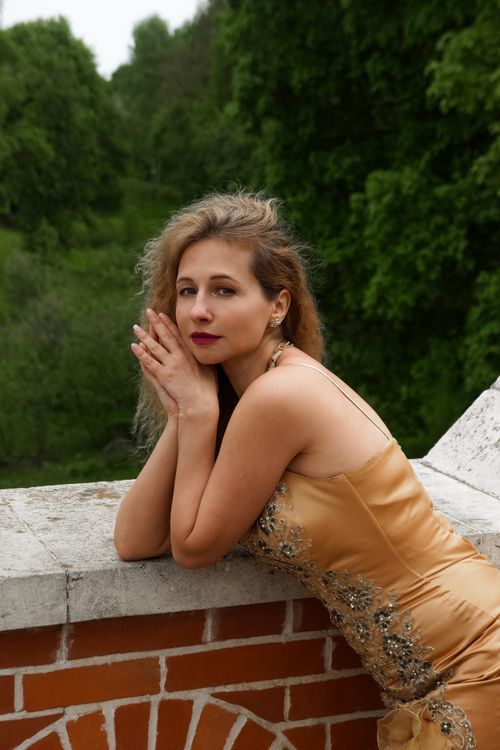 Free A woman in a gold dress poses gracefully against a brick wall in a lush outdoor setting. Stock Photo