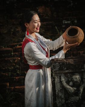 Woman in traditional attire holding an earthen pot against a rustic brick background.