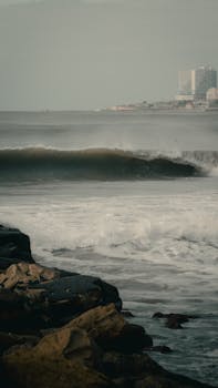 Dramatic ocean waves hitting the rocky shore of Mar del Plata with cityscape in the background.