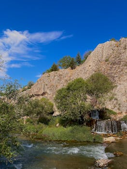 Beautiful landscape showing a flowing river, waterfall, and rocky cliff under a clear blue sky.