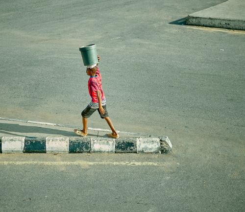 Free A child in a pink shirt carries a bucket on head while walking on urban pavement. Stock Photo