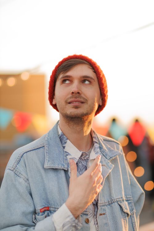 Free A young man wearing a red beanie and denim jacket looks upwards with bokeh lights behind him during sunset. Stock Photo