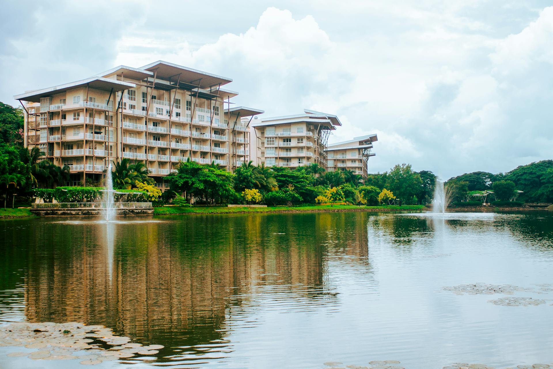 Complex of modern buildings located on shore of rippling lake with fountains surrounded by lush green trees against cloudy sky in Philippines