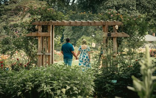 Free Romantic couple holding hands and walking through a beautiful garden archway, surrounded by lush greenery and vibrant flowers. Stock Photo