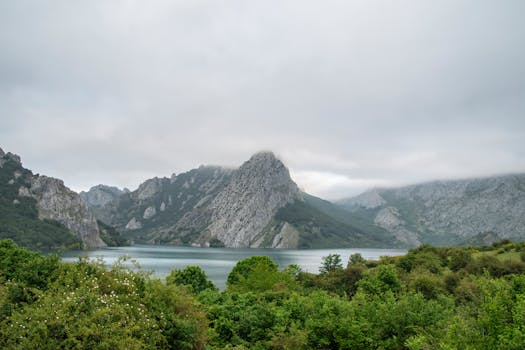 Serene mountain lake surrounded by lush greenery and misty peaks under cloudy sky.