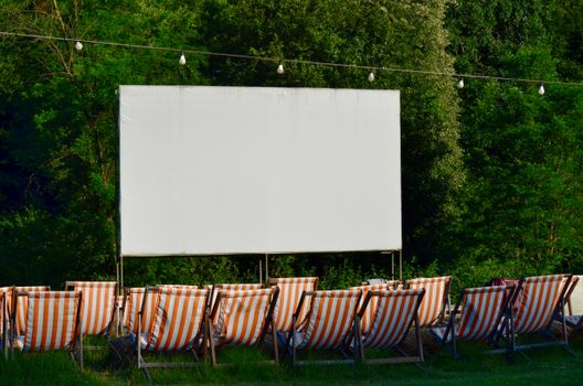 Empty outdoor cinema venue with striped deck chairs and a blank screen, surrounded by greenery.