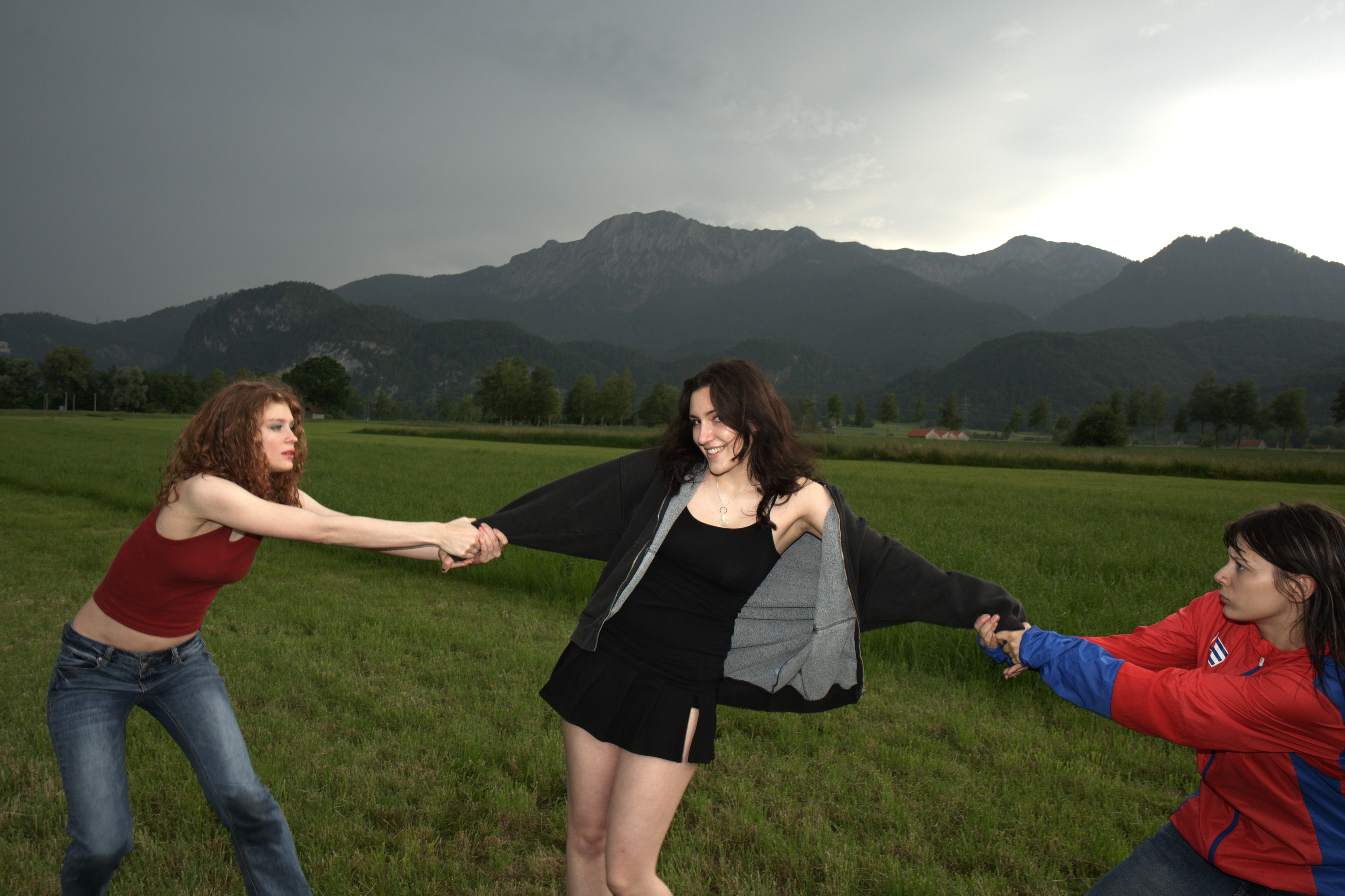 Three friends enjoy playful tugging in a lush green field with mountains.