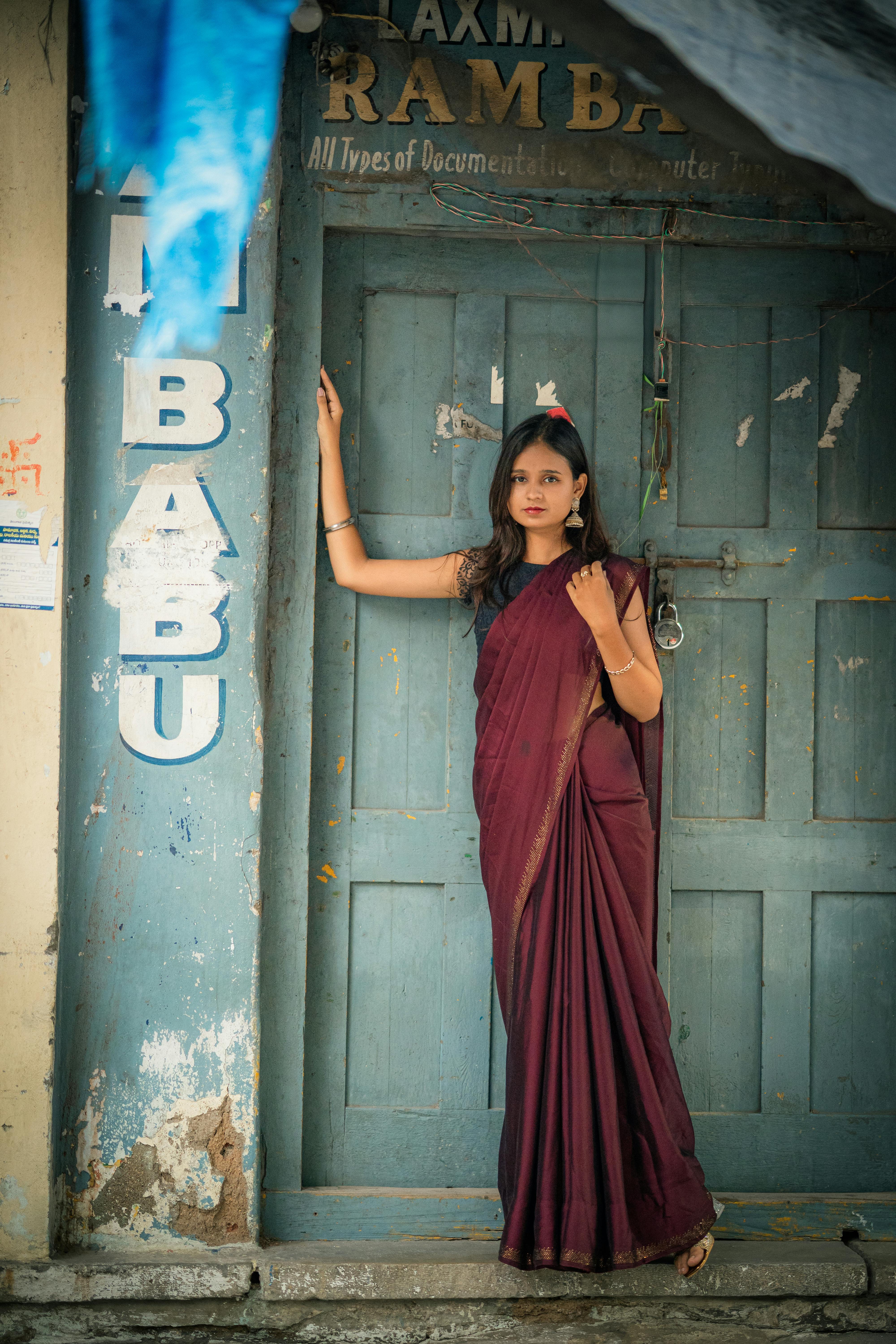 Woman in a burgundy saree stands gracefully against a rustic wooden door in an urban setting.