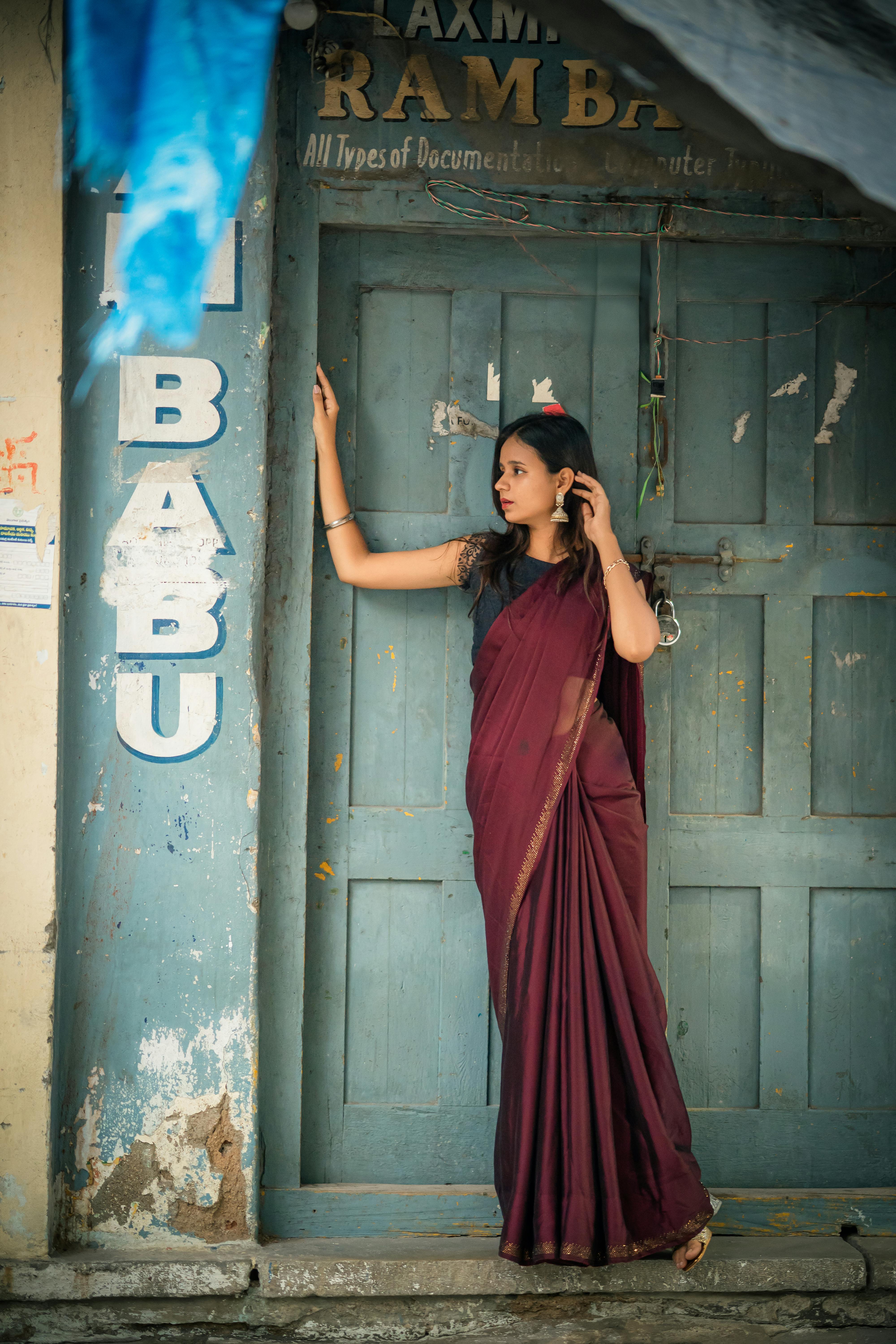 A stylish woman in a burgundy saree poses thoughtfully against a vintage door with weathered paint.