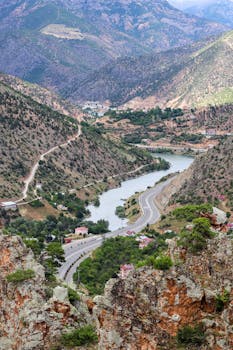 Aerial view of a winding road through mountains with a river bend and lush green landscape.