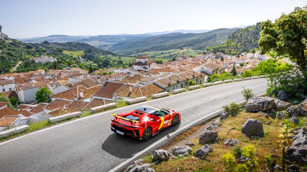 A vibrant red sports car races through a scenic Spanish village with mountains in the background.