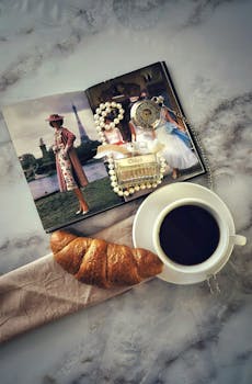 Flat lay of a Parisian breakfast with croissant, coffee, vintage magazine, jewelry, and pocket watch on a marble surface.