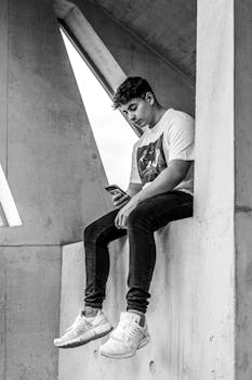 Stylish young man sitting indoors, engaged with smartphone in a monochromatic setting.