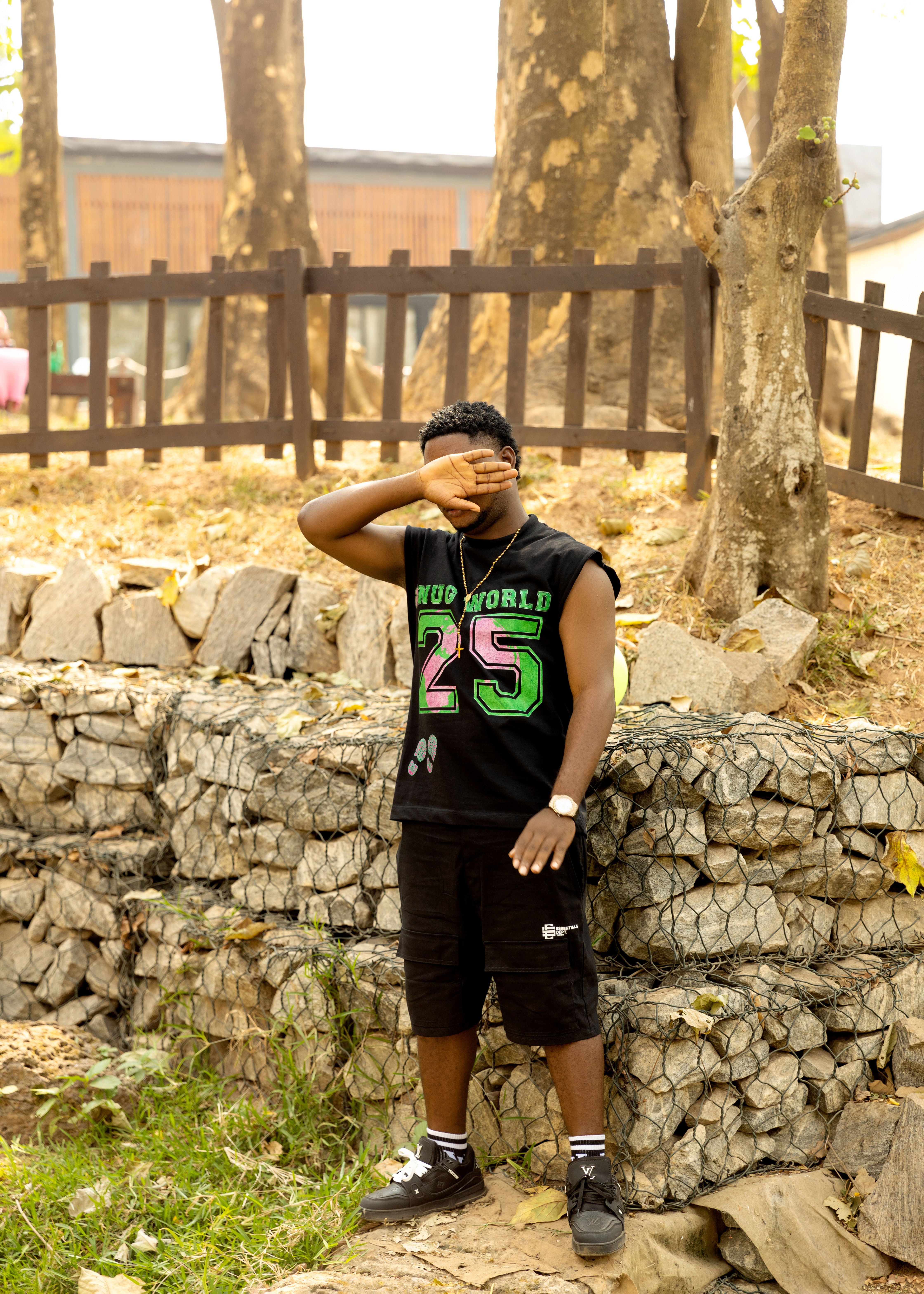 A teenager poses casually outdoors in a summer outfit, covering face with hand.