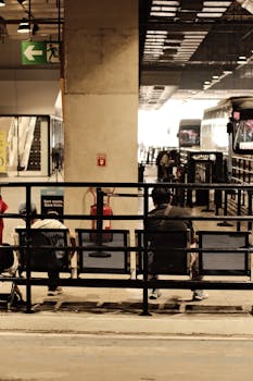 Passengers waiting at a bus station with visible signs and buses in the background.