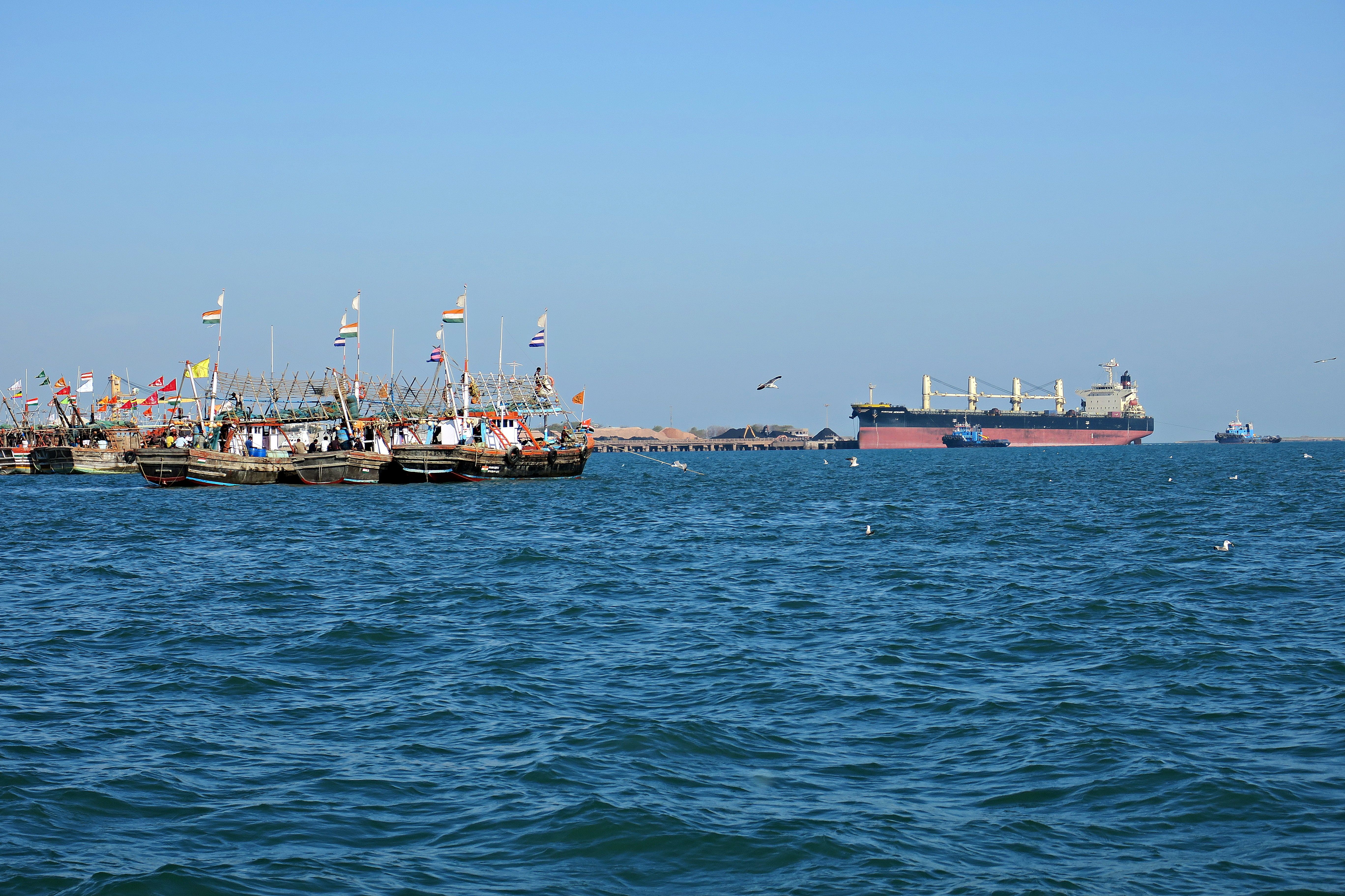 A scenic view of colorful fishing boats with flags and a cargo ship on the ocean.