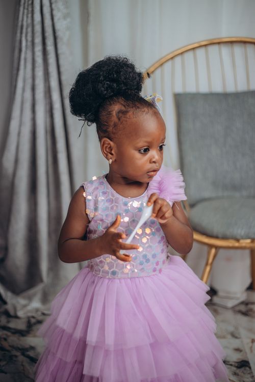 Free Cute toddler in a lavender dress standing indoors with a playful expression. Stock Photo