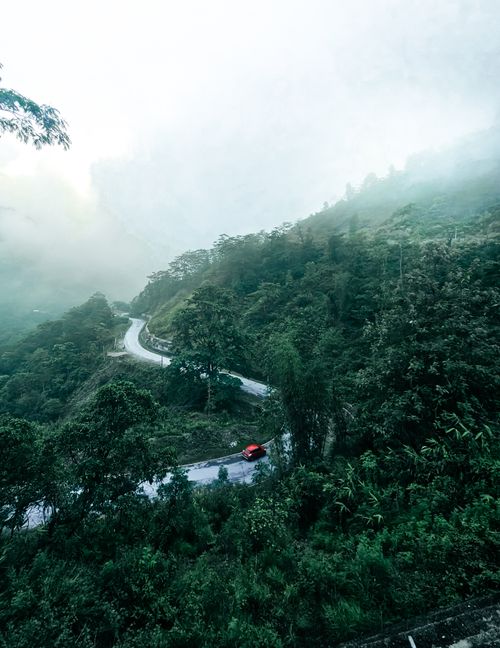 Free A scenic view of a winding mountain road enveloped in fog with a red car driving through lush greenery. Stock Photo