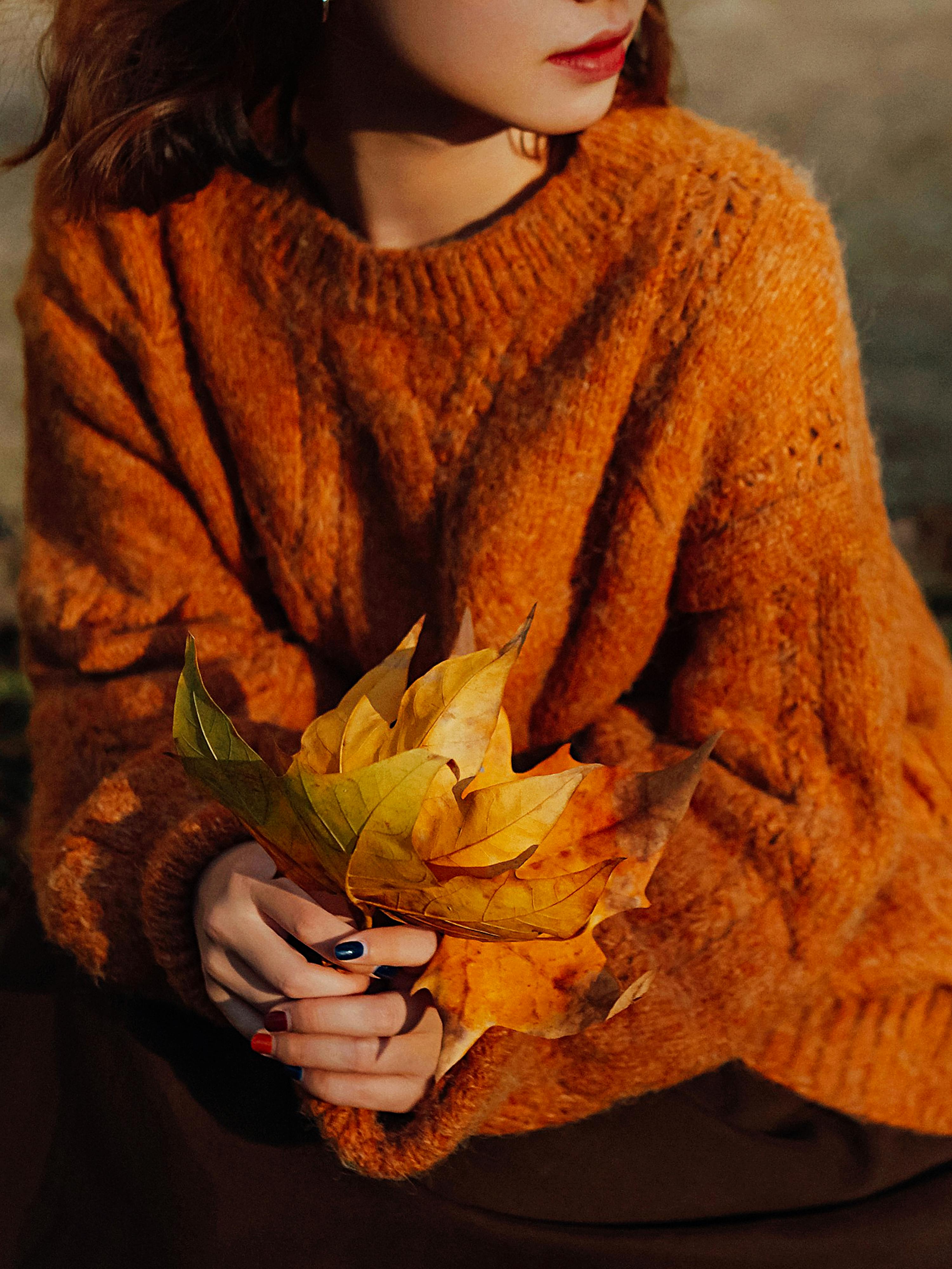 A woman in an orange sweater holding autumn leaves, capturing a warm fall moment.
