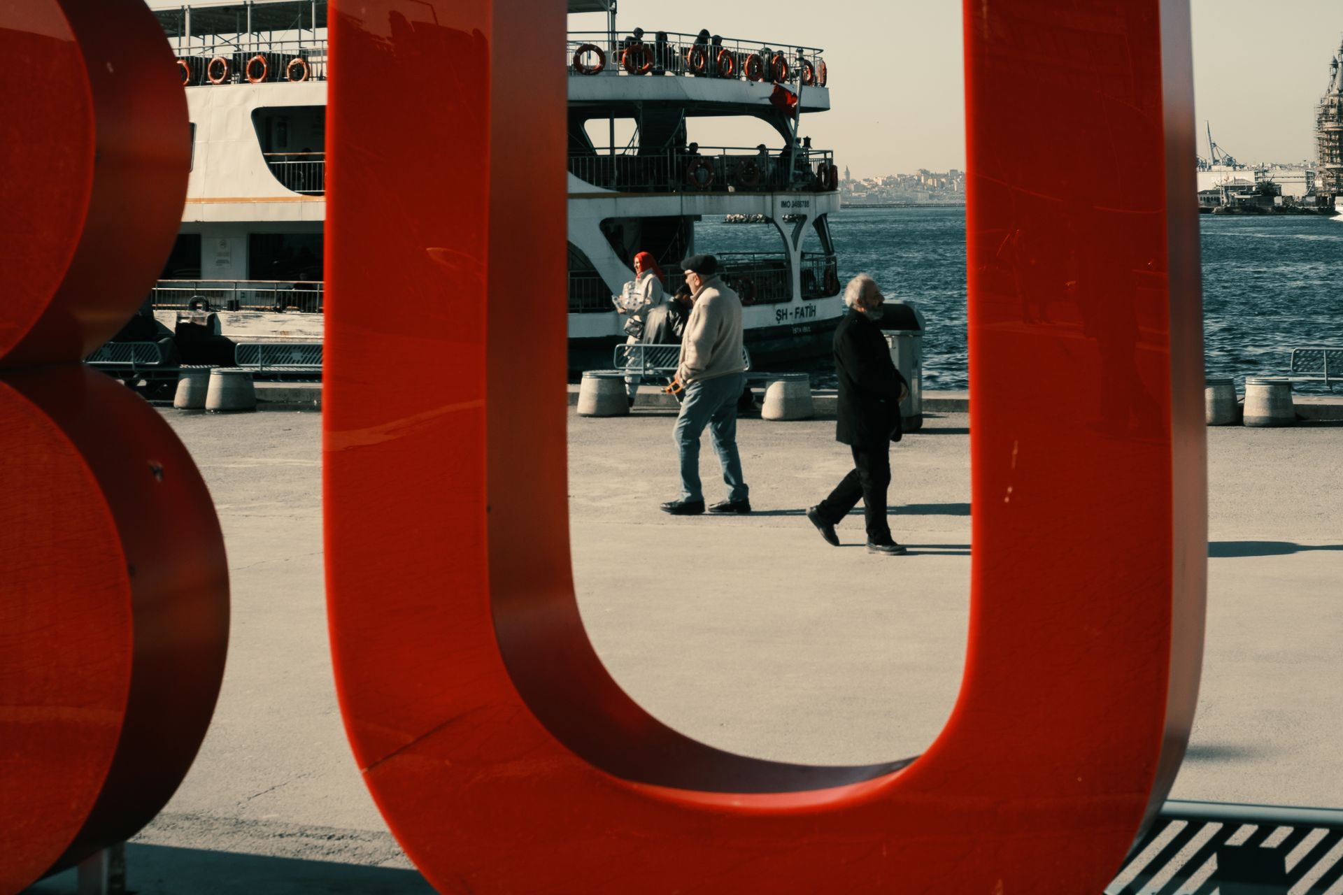 Scene of pedestrians passing large red sculpture near a ferry terminal with cityscape background.