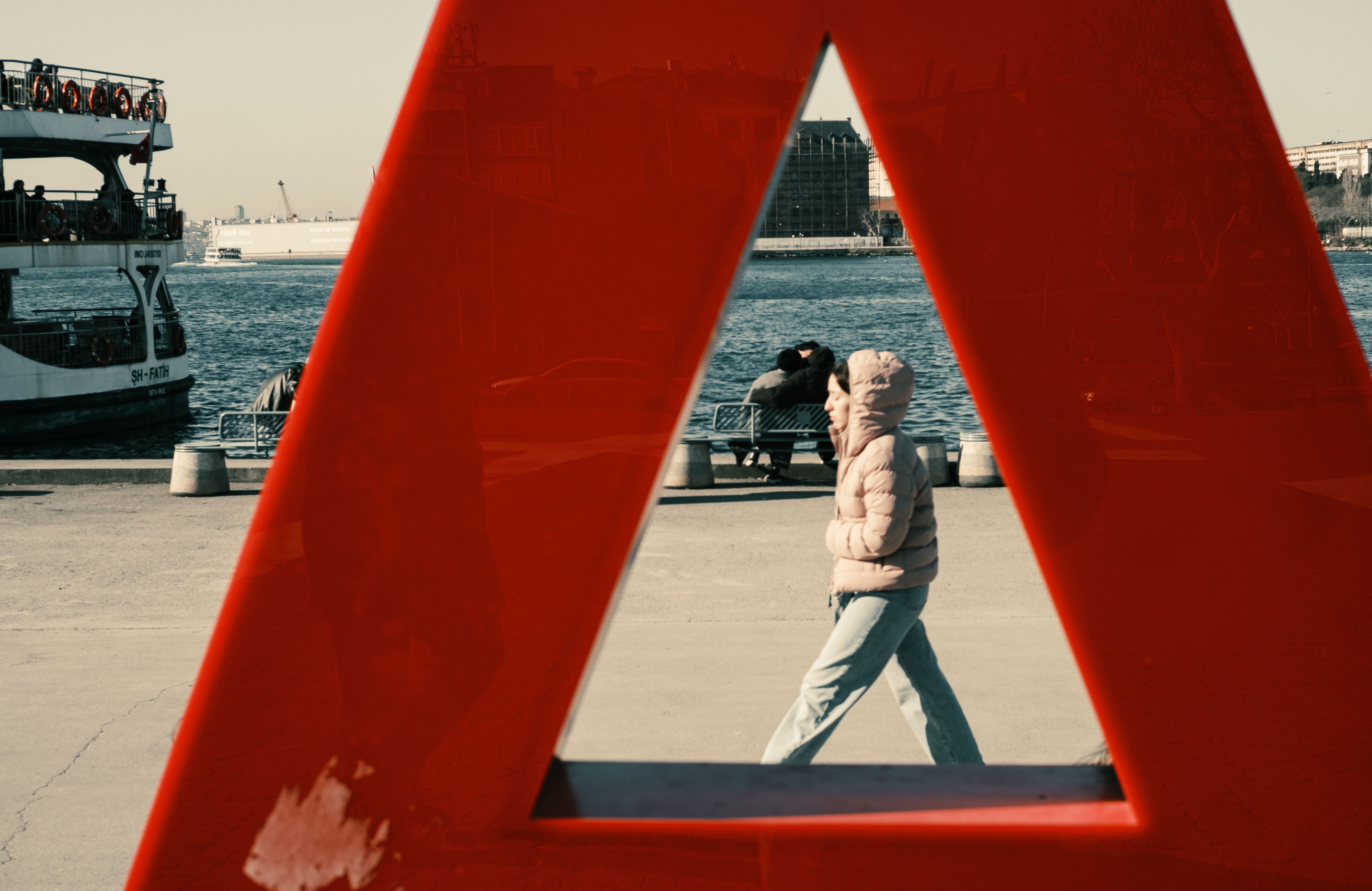 A person walks by waterfront framed through a red sculpture with a ferry in the background.