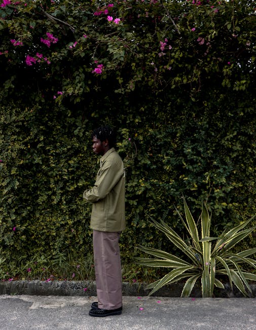 A man in casual streetwear poses against a backdrop of lush green foliage and bright flowers, embodying street fashion.