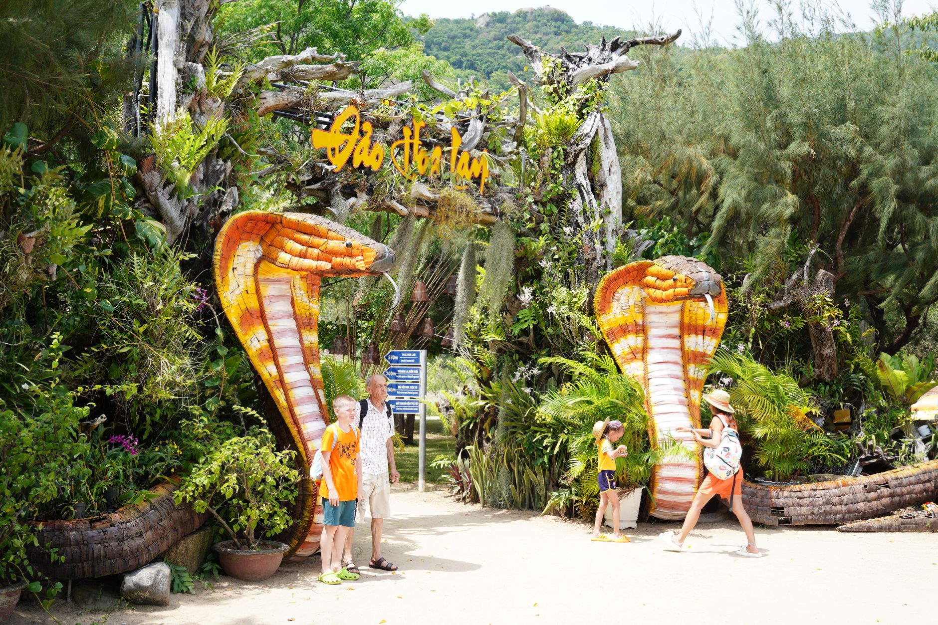 Tourists admire colorful snake sculptures at a tropical outdoor attraction, surrounded by lush greenery.