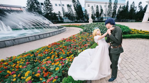 Cosplay couple in colorful garden with fountain backdrop, outdoor setting.