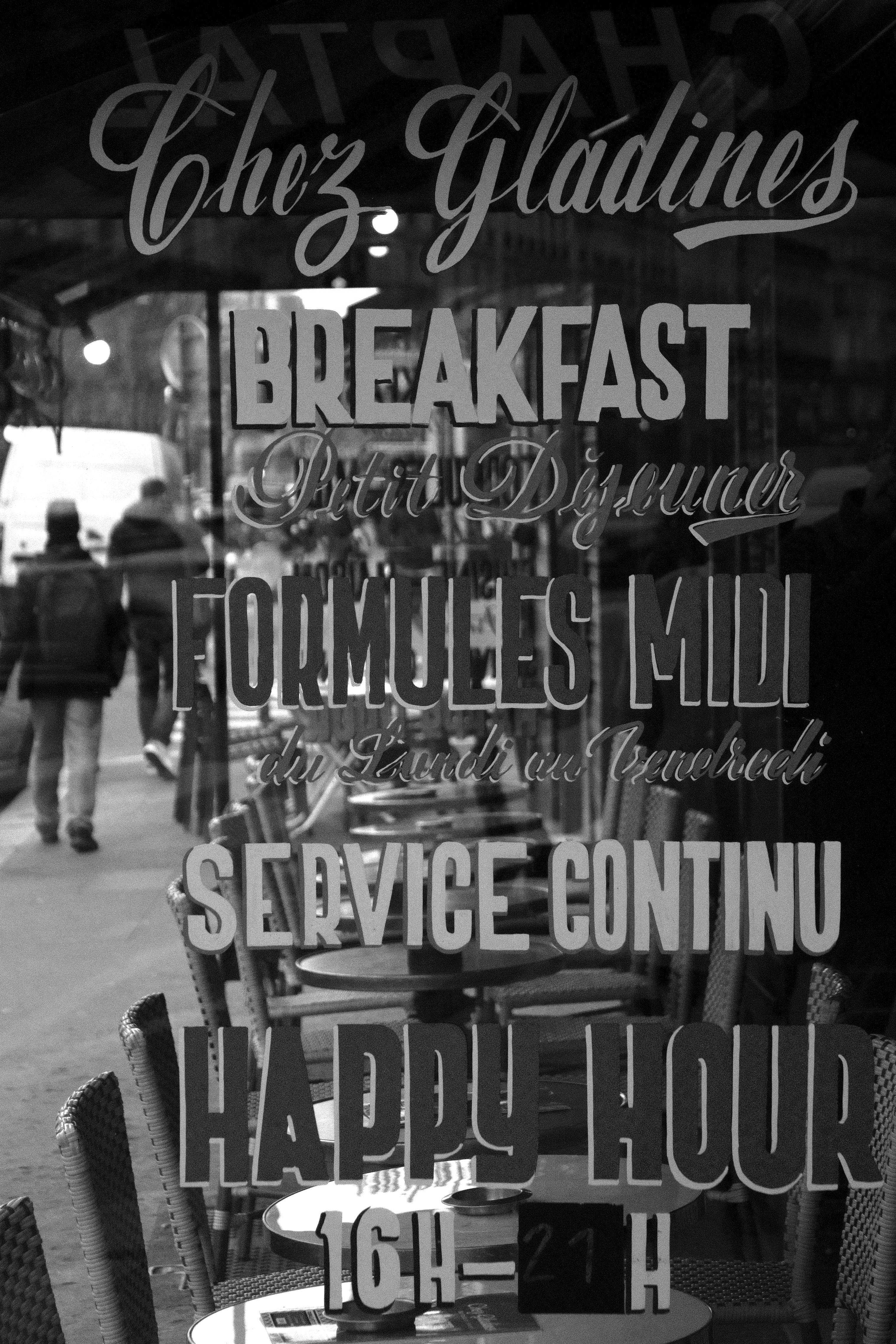 Black and white photo capturing reflective French cafe signage in Paris. People walking by.