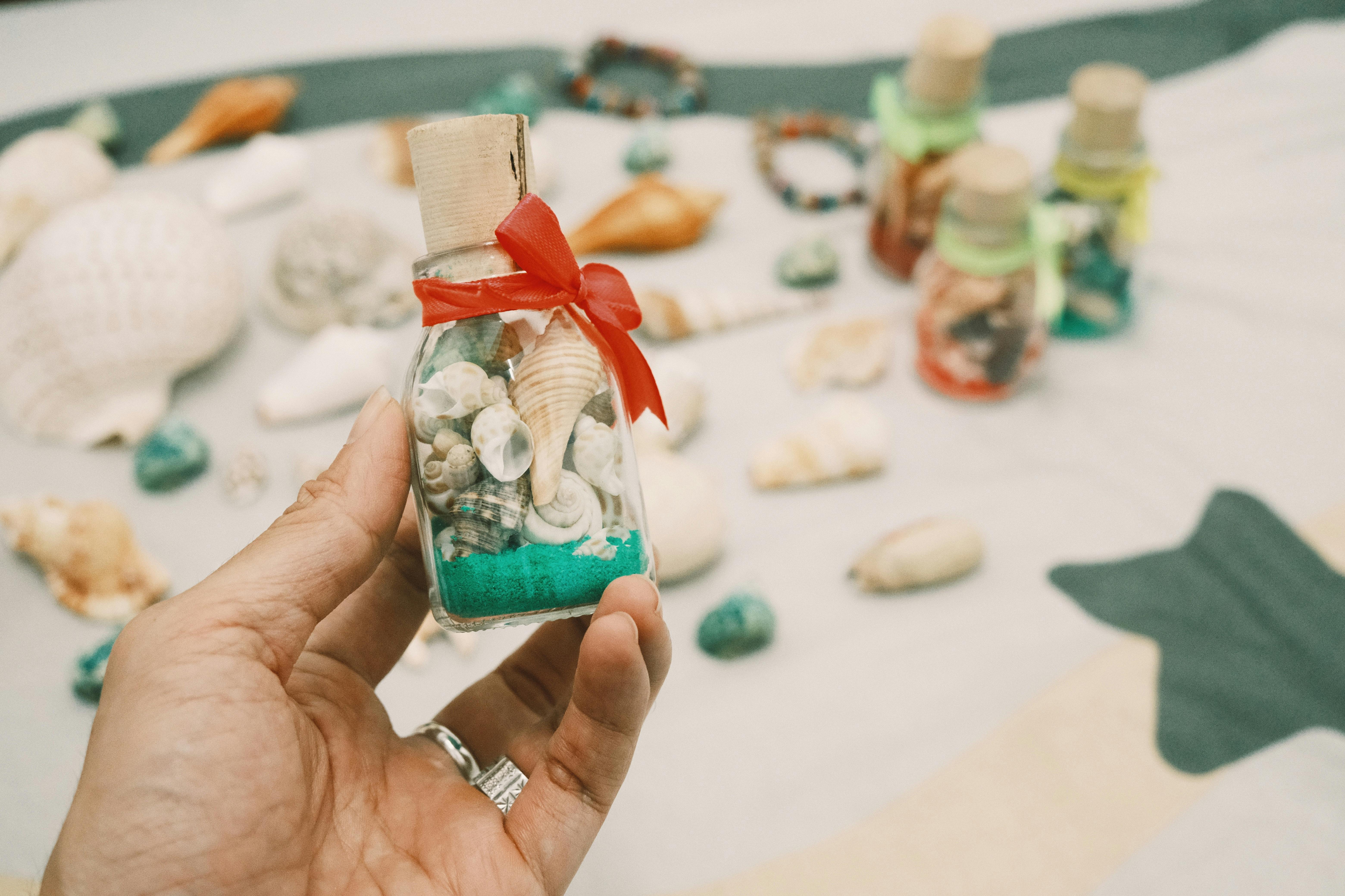 Close-up of a hand holding a decorative bottle filled with shells and sand, set on a beach-inspired background.