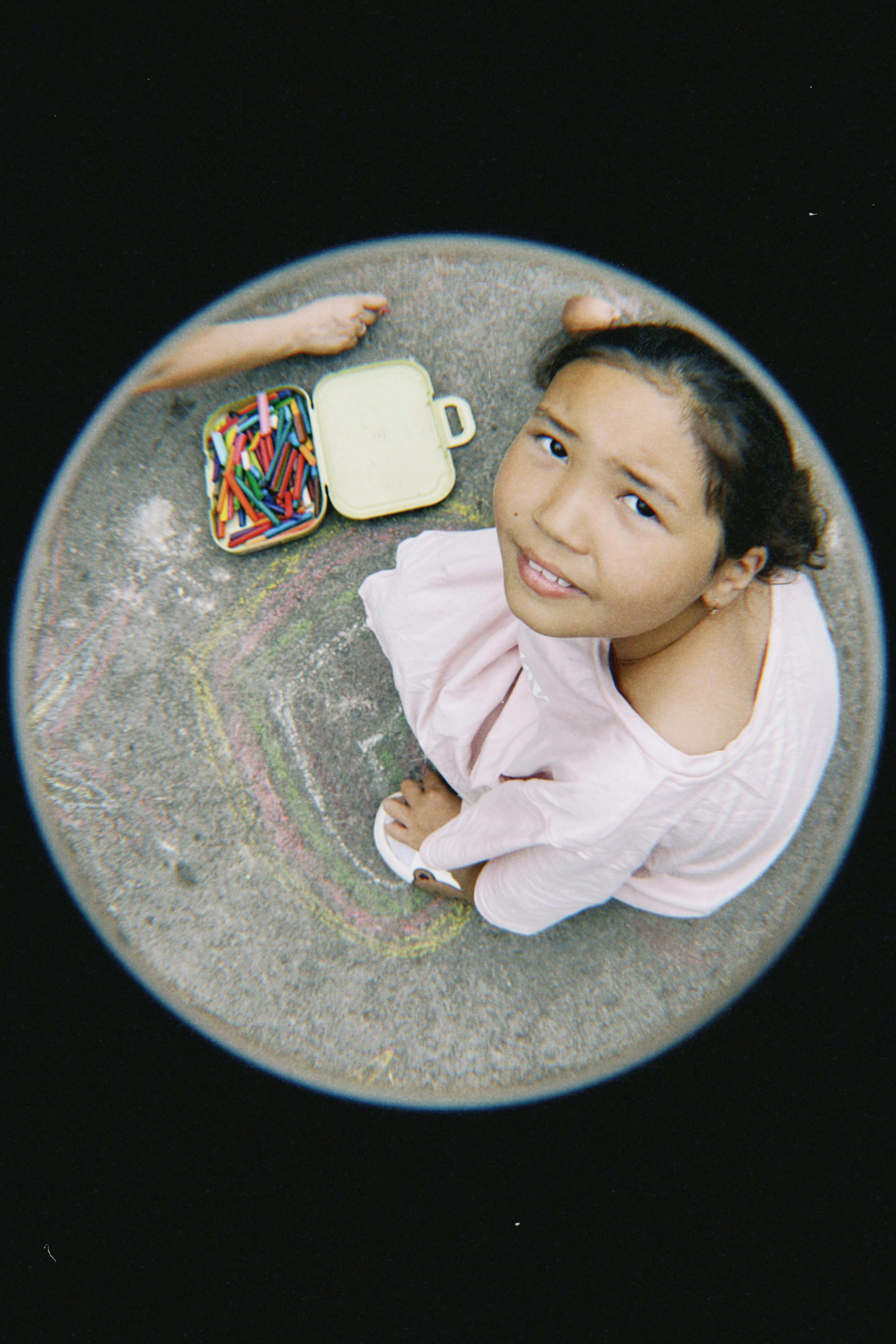 A child with chalks creating art on the pavement, captured through a fisheye lens.