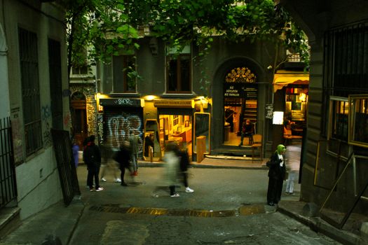 Blurred pedestrians move through an illuminated urban alley at night, capturing the lively street atmosphere.