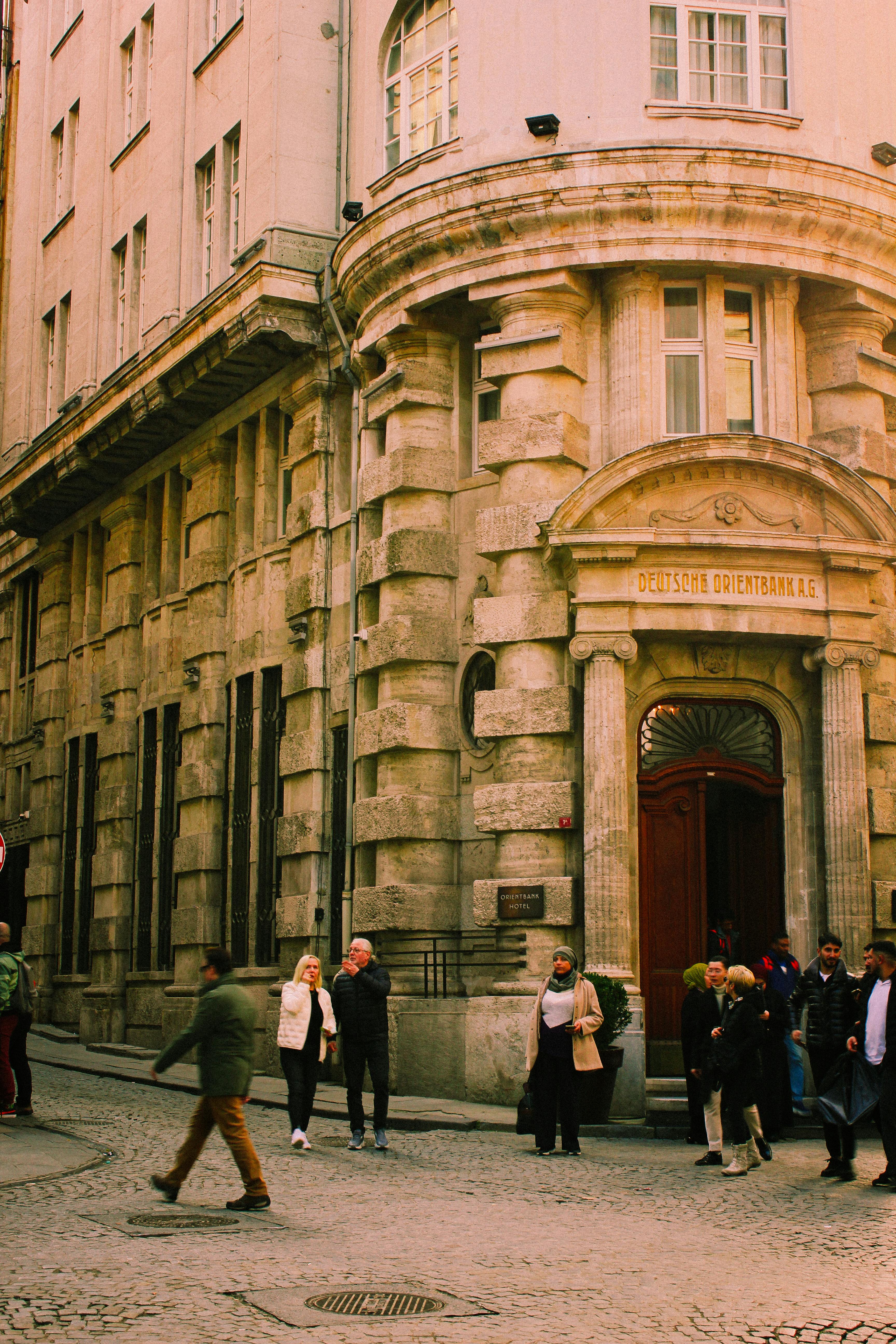 Kostenlos Menschen, die in einer städtischen Umgebung an einem historischen Bankgebäude auf einer Kopfsteinpflasterstraße vorbeigehen. Stock-Foto