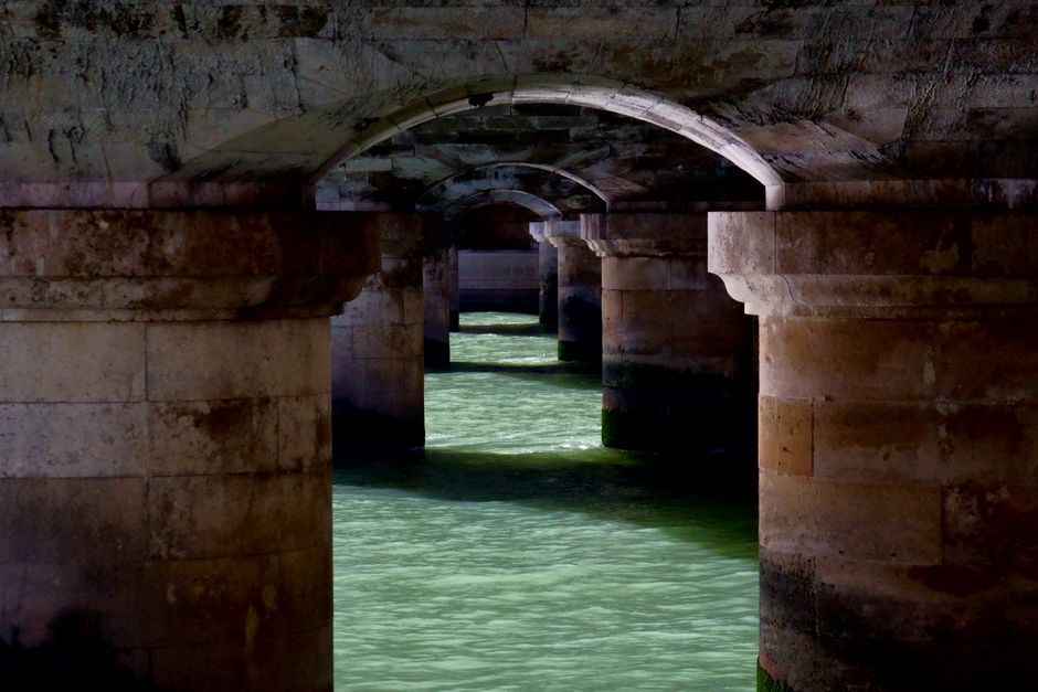 View under a Paris bridge showing stone arches reflecting on the Seine River.