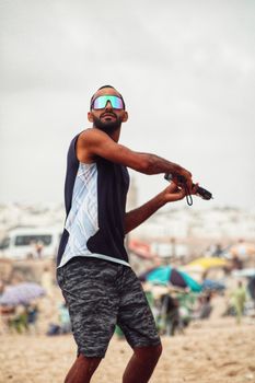 A man with sunglasses and a tank top plays frisbee on a lively beach.