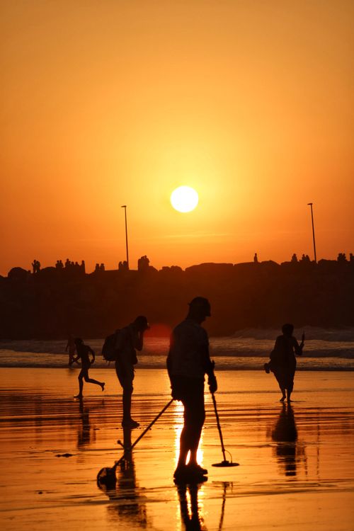 Free Silhouetted people using metal detectors along a sunlit beach during a vivid sunset, creating a scenic and serene atmosphere. Stock Photo
