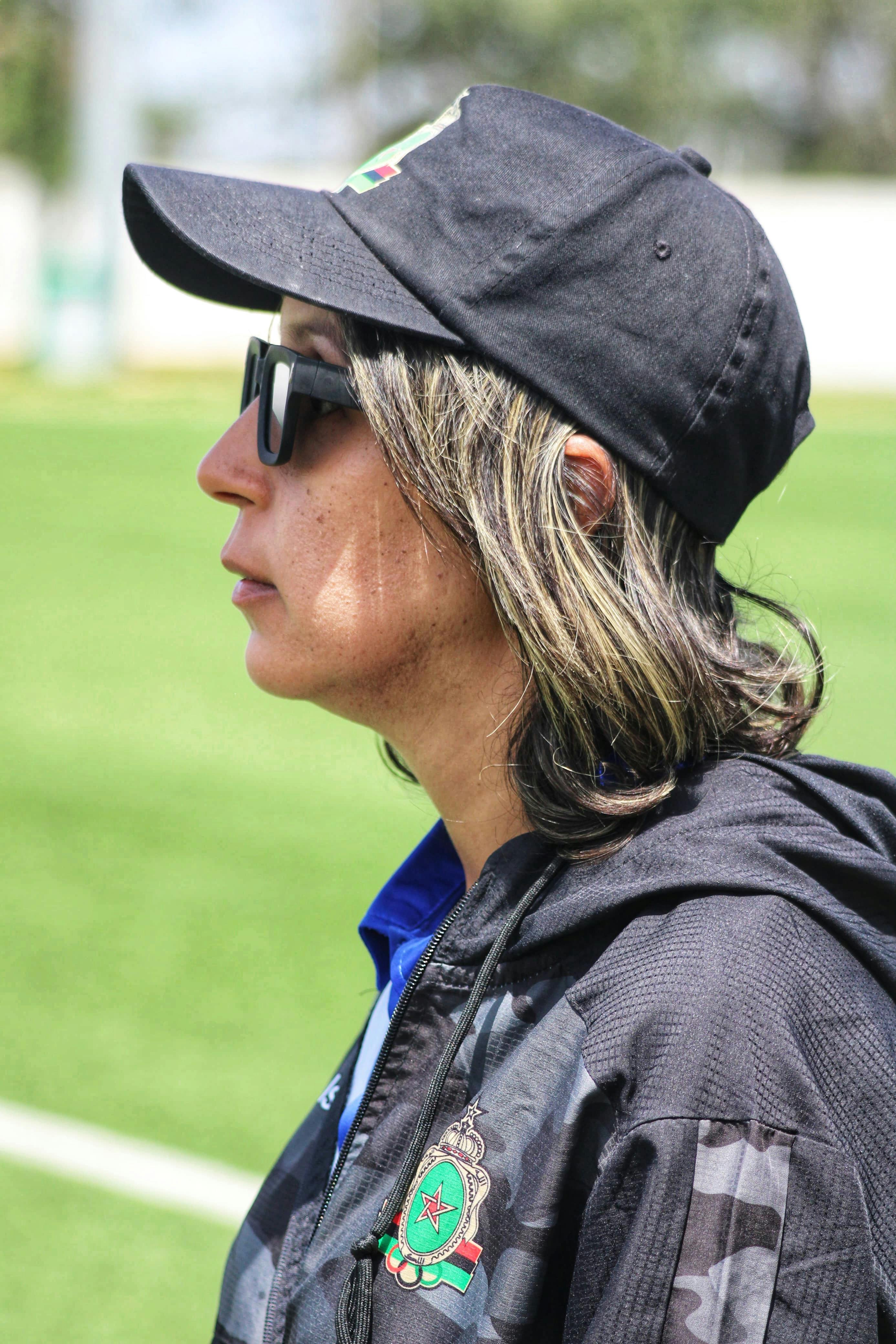 Side view of a woman in sunglasses and black cap on a sunny sports field.