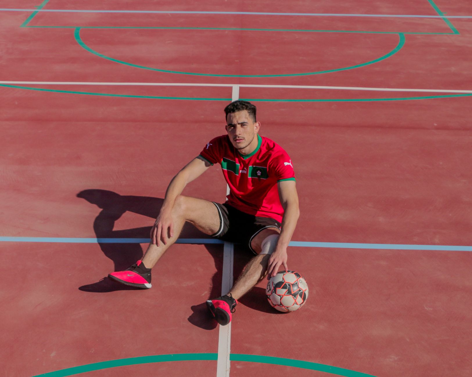 Athletic man in sports attire sitting on a red outdoor court with a soccer ball.