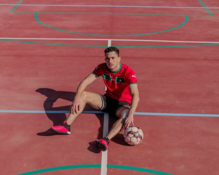 Athletic man in sports attire sitting on a red outdoor court with a soccer ball.
