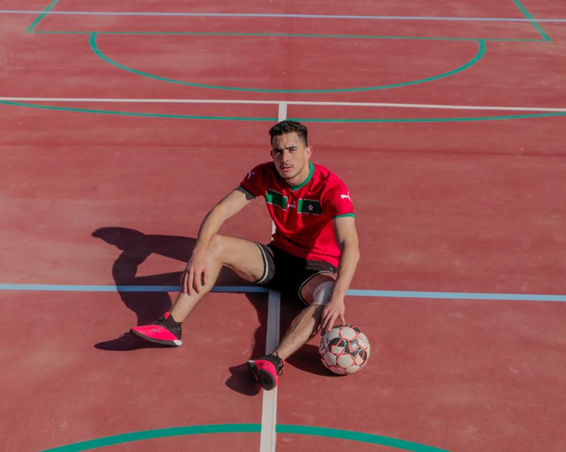 Athletic man in sports attire sitting on a red outdoor court with a soccer ball.