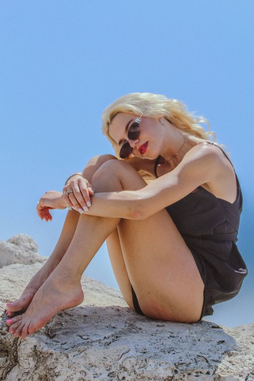 Free A woman sitting on rocks in a black dress and sunglasses, enjoying a sunny day. Stock Photo