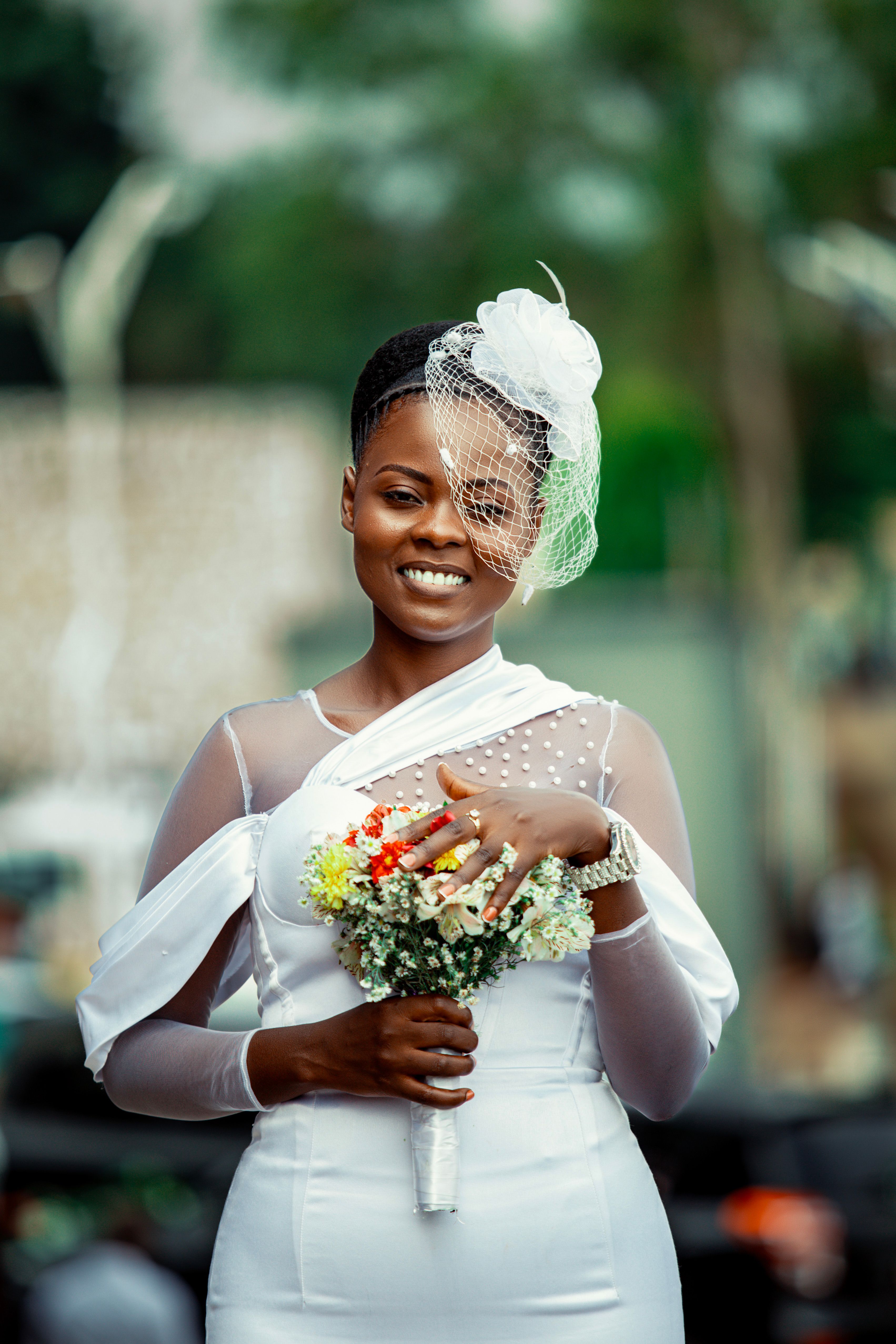 Beautiful black bride smiling outdoors in white dress. She holds a bouquet and wears a stylish fascinator.