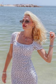 Blonde woman in sunglasses and floral dress enjoying a sunny day by the beach.