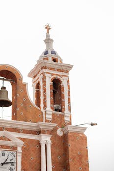 A detailed view of a historic church tower with a clock, bell, and cross against a clear sky.