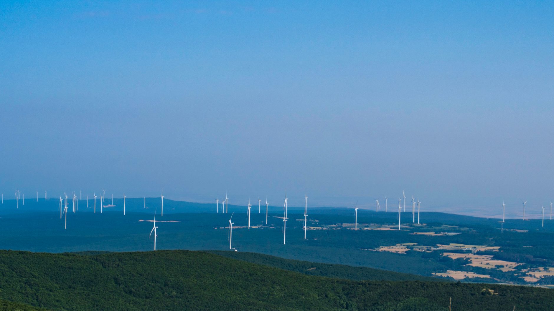 Expansive wind farm with multiple turbines on a green hilly terrain under a clear blue sky.