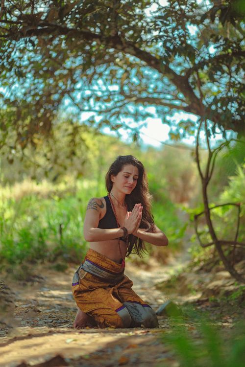 Free A woman kneels in a meditative pose under a tree in a sunny outdoor setting. Stock Photo