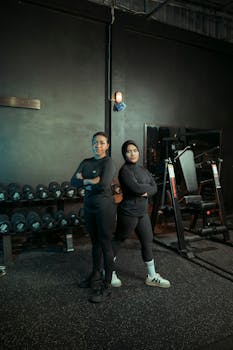 Two women in sportswear stand confidently in a gym with equipment in the background, promoting fitness and empowerment.