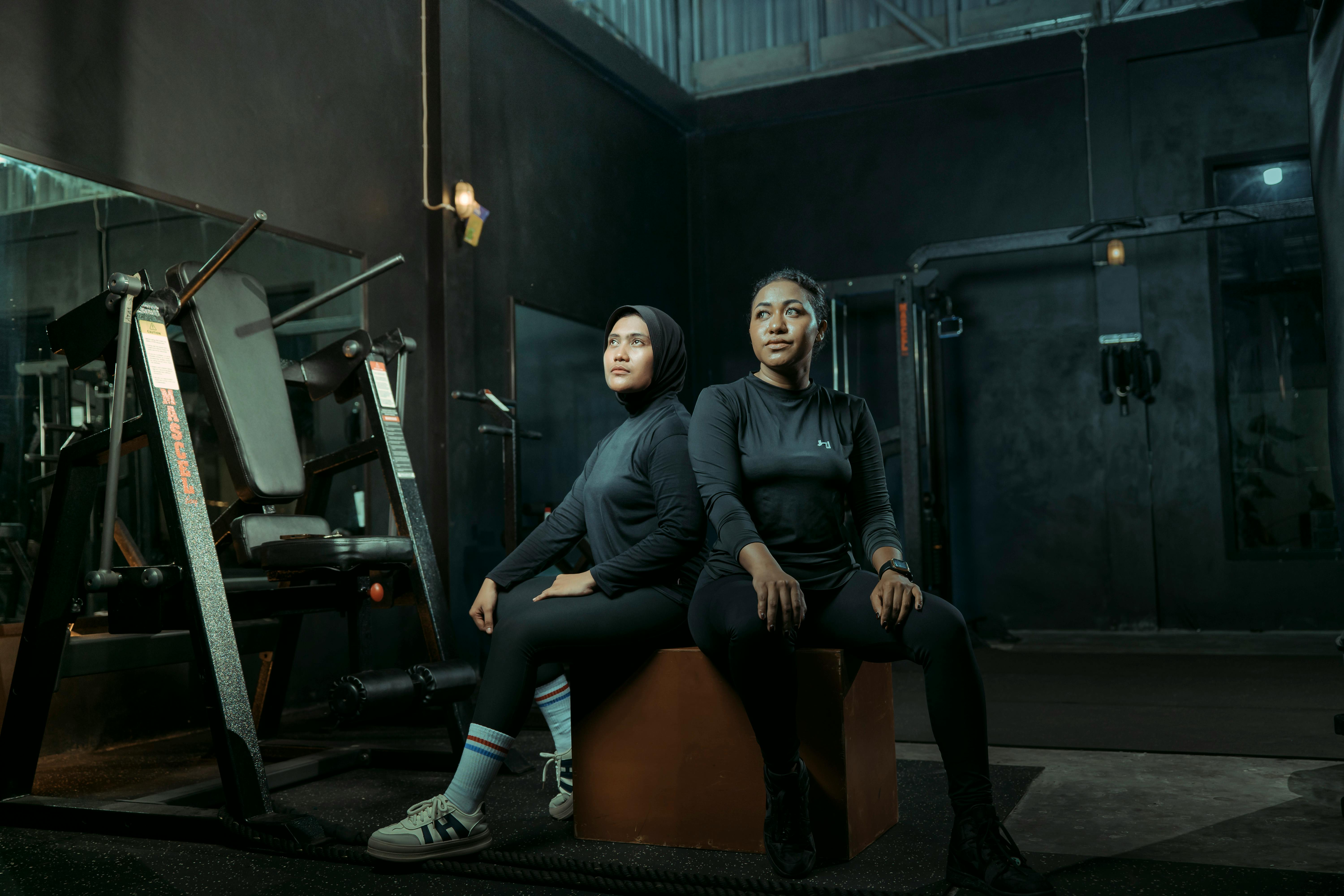 Two women in athletic wear sitting on a box with gym equipment in the background.
