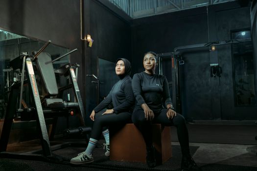 Two women in athletic wear sitting on a box with gym equipment in the background.
